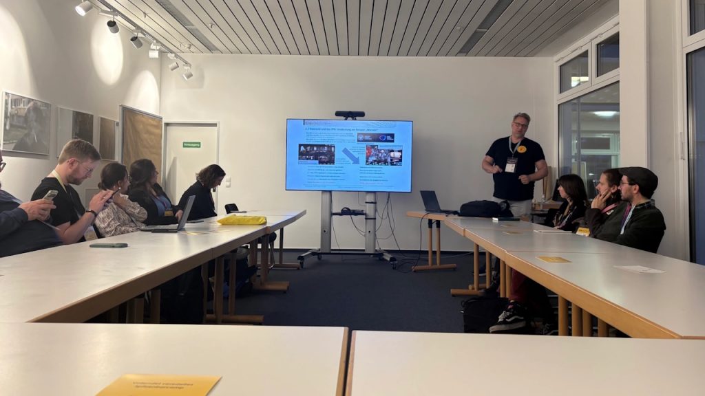 Personen sitzen in einem Konferenzraum bei einer Session auf dem histocamp 26. Mittig der Monitor mit einer Präsentation, rechts Nolden mit seinem Impuls