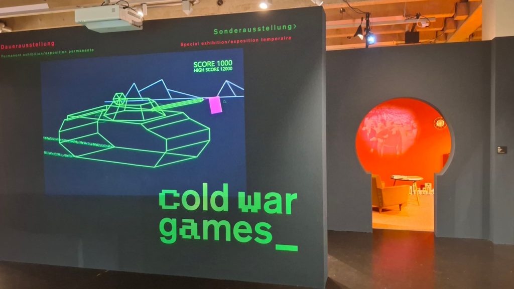 Links projeziert ein Beamer auf die Ausstellungswand einen Panzer in stilisierter Vektorgrafik. Unten zu sehen: der Titel der Ausstellung. Rechts schimmert der Raum mit Möbeln und Informationen aus den fünfziger Jahren durch eine Tür, die wie ein Schlüsselloch erscheint.