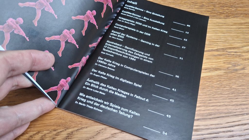 Links im Bild meine Hand und darüber in rosa kleine Plastiksoldaten, deren Waffen auf die enthaltenen Fachbeiträge des Kataloges zeigen. Im Hintegrund die hölzerne Ablage meines Schreibstischs.