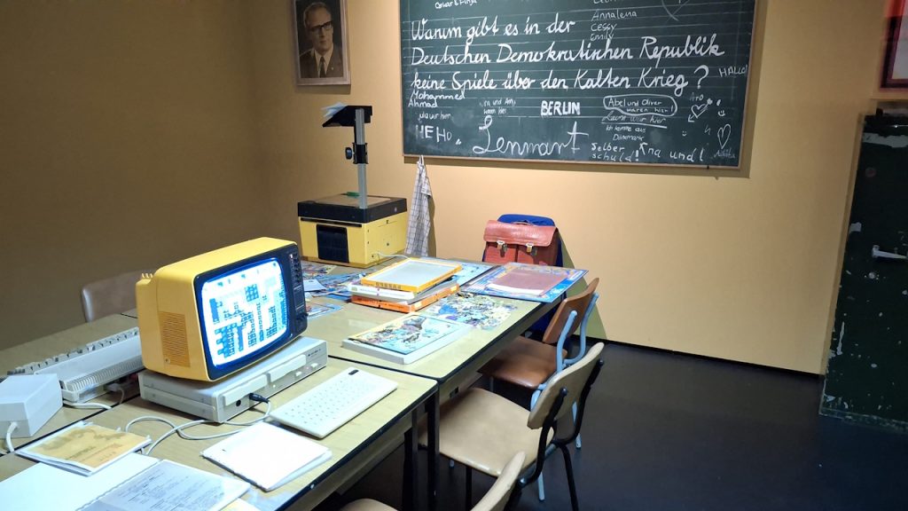 Im Hintergrund ein Portrait des Staatsratsvorsitzenden Erich Honecker aus der DDR mit einer Frage auf der Schiefertafel: "Warum gibt es in der DDR keine Spiele über den Kalten Krieg?" Davor digitale und analoge Spiele zum Thema aus den achtziger Jahren mit einem TV-Gerät und einer Konsole. 